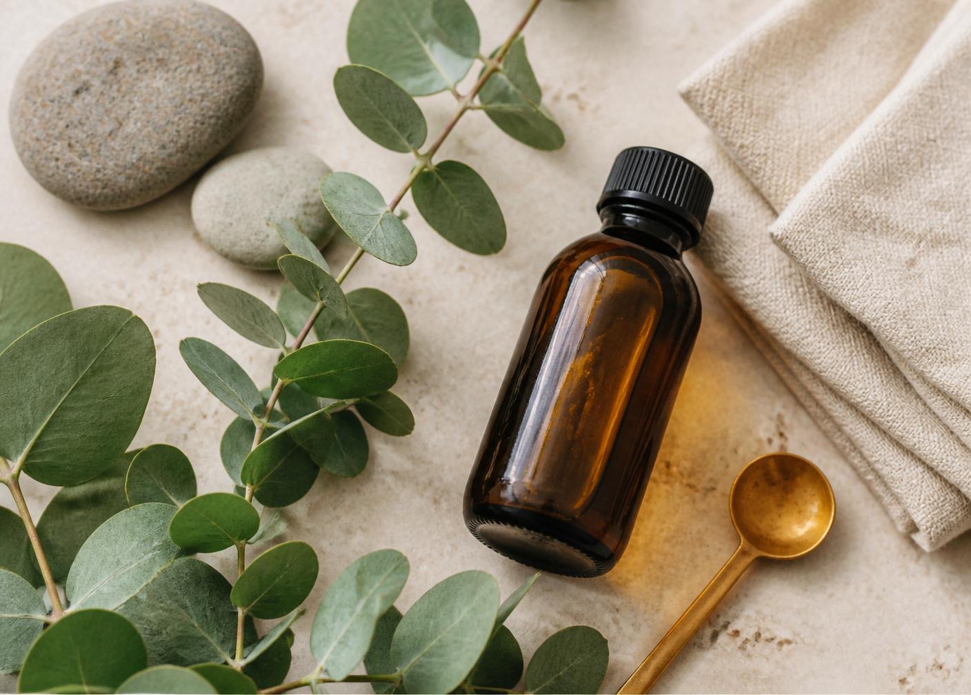 Flat lay of eucalyptus branches, smooth river stones, an amber oil bottle, folded linen, and a brass spoon